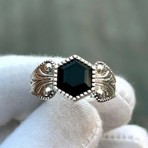 Op de afbeelding: Een zilveren ring met een grote, zeshoekige zwarte edelsteen. De ring heeft gedetailleerde krullen aan de zijkanten van de band. De edelsteen is gezet in een bezel setting met een gestippelde rand.