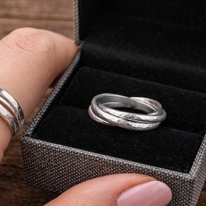 May include: A silver ring with a hammered texture is displayed on a finger and in a gray ring box. The ring has three intertwined bands. The ring box has a black interior and is open, revealing the ring.