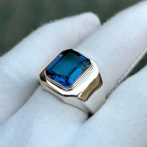 Peut inclure: Une bague en argent avec une grande pierre précieuse rectangulaire bleu foncé. La pierre est sertie dans un anneau en argent poli. La bague est présentée sur une surface blanche.