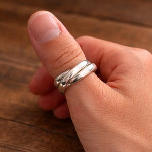 Könnte beinhalten: Ein silberner Ring mit einem verdrehten Design. Der Ring wird an einem Daumen getragen. Der Ring hat eine polierte Oberfläche und reflektiert das Licht. Der Ring scheint aus Metall zu sein.