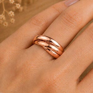 Puede incluir: Un anillo de oro rosa con un diseño de tres bandas con superficies lisas y pulidas. Las tres bandas interconectadas están entrelazadas para crear una pieza de joyería cohesiva que se asienta sobre el dedo con un perfil redondeado.