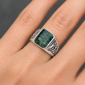 May include: A silver ring with a rectangular, emerald-green gemstone. The ring has an ornate, engraved design on the band. The gemstone is set in a bezel setting, and the ring is shown on a finger.