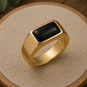 May include: A gold-toned signet ring featuring a smooth, rectangular black inlay set into the center of the flat top. The band is polished and solid, with a thick, rounded profile that tapers slightly toward the bottom of the ring's circular shank.