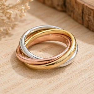 Op de afbeelding: Een driekleurige rolling ring bestaande uit drie in elkaar grijpende banden in gepolijste zilver-, geelgoud- en roségoudtinten. De gladde, afgeronde metalen banden zijn met elkaar verbonden tot één sieraad met een reflecterende afwerking.