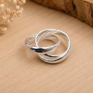 Può includere: Un anello color argento lucido composto da tre fasce intrecciate che si sovrappongono per formare un'unità circolare. La superficie metallica è liscia e riflettente, catturando la luce per evidenziare la forma arrotondata di ogni singola fascia.