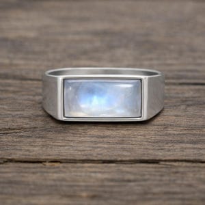 Puede incluir: Anillo de plata con una piedra lunar rectangular e iridiscente. La piedra lunar tiene un suave brillo azul y blanco. El anillo tiene una banda gruesa y rectangular. Sobre un fondo de madera.
