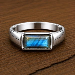 May include: A silver ring with a rectangular, iridescent blue gemstone. The ring has a polished finish and a simple, modern design. The gemstone displays shades of blue, green, and yellow.
