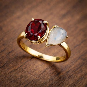 Puede incluir: Un anillo de oro con una gema grande, redonda y de color rojo intenso y una gema más pequeña, blanca y en forma de pera. El anillo se exhibe sobre una superficie de madera, resaltando el diseño y el contraste de color.
