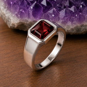 May include: A silver-toned ring featuring a large, rectangular, deep red gemstone set in a smooth, polished metal band. The setting is geometric and clean, highlighting the faceted cut of the dark red stone which sits prominently on the band.