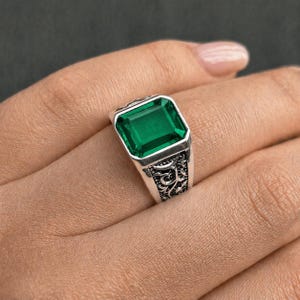 May include: A silver ring with a large, emerald-cut green gemstone. The ring has an ornate, engraved design on the band. The gemstone is rectangular and set in a silver bezel.