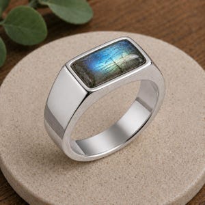May include: A silver-toned signet ring featuring a rectangular, polished labradorite gemstone inlay. The stone displays a vibrant blue and green iridescent flash across its surface, set within a smooth, solid metal band with a reflective finish.