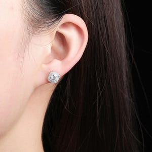 May include: A close-up of a square-shaped stud earring with a halo of small, clear stones surrounding a larger, clear stone. The earring is set in a silver-colored metal and is worn on an ear.