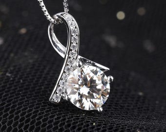 May include: A silver pendant necklace featuring a large, round, clear gemstone. The pendant has a curved, ribbon-like design, embellished with smaller, clear gemstones. The necklace chain is a delicate silver. The background is a dark, textured fabric.