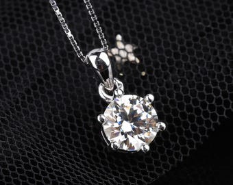 May include: A silver pendant necklace featuring a large, round, clear gemstone. The gemstone is held in place by six prongs. The chain is a delicate silver link style, and the background is a dark, textured fabric.