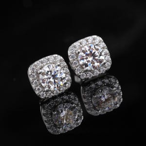 May include: A pair of square-shaped stud earrings with a large, round, clear center stone surrounded by smaller, clear stones. The earrings are set in a silver-colored metal and reflect light, creating a sparkling effect. The background is black.