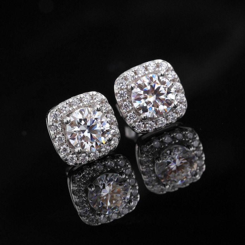 May include: A pair of square-shaped stud earrings with a large, round, clear center stone surrounded by smaller, clear stones. The earrings are set in a silver-colored metal and reflect light, creating a sparkling effect. The background is black.