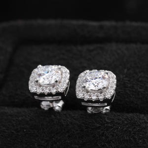 May include: A pair of silver stud earrings. Each earring features a large, round, clear stone surrounded by a square halo of smaller, clear stones. The earrings are set against a black background.