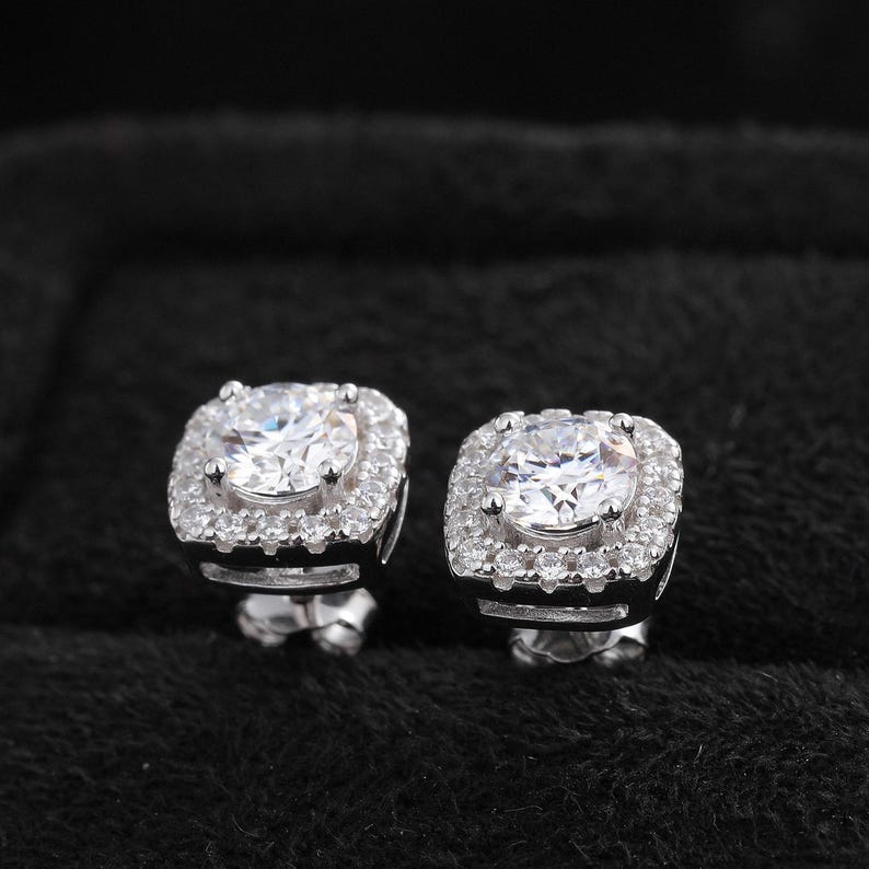 May include: A pair of silver stud earrings. Each earring features a large, round, clear stone surrounded by a square halo of smaller, clear stones. The earrings are set against a black background.