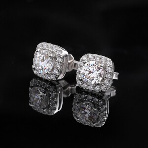 May include: A pair of square-shaped stud earrings with a large, clear, round center stone surrounded by smaller stones. The earrings are set in a silver-colored metal and reflect on a black surface.