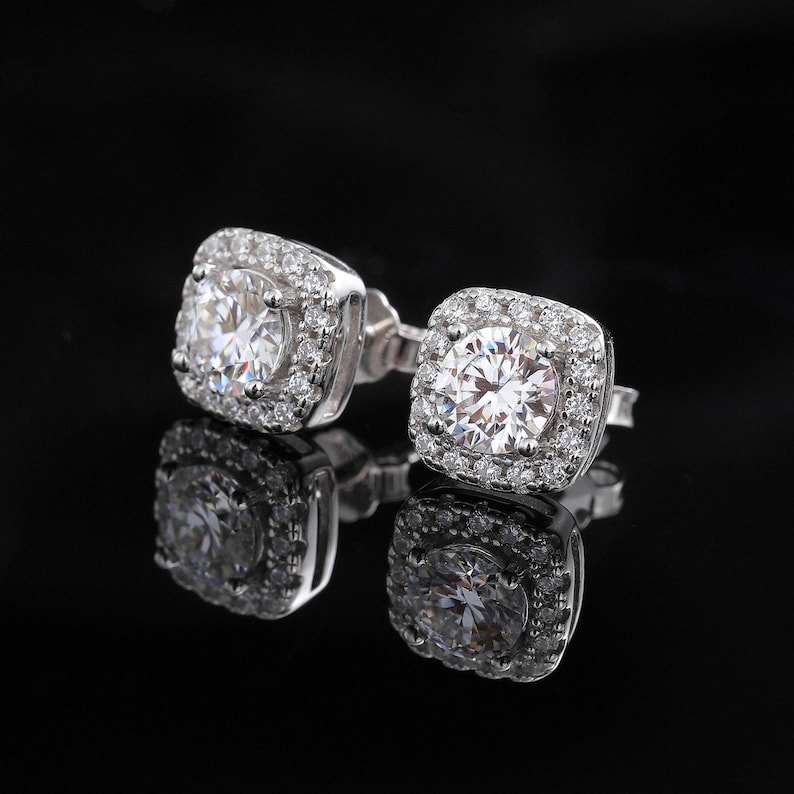 May include: A pair of square-shaped stud earrings with a large, clear, round center stone surrounded by smaller stones. The earrings are set in a silver-colored metal and reflect on a black surface.