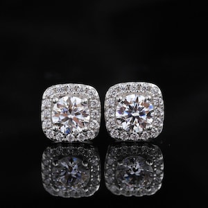 May include: A pair of square-shaped, silver-toned stud earrings. Each earring features a large, round, clear gemstone surrounded by smaller, clear gemstones. The earrings are set against a black background, with their reflections visible.