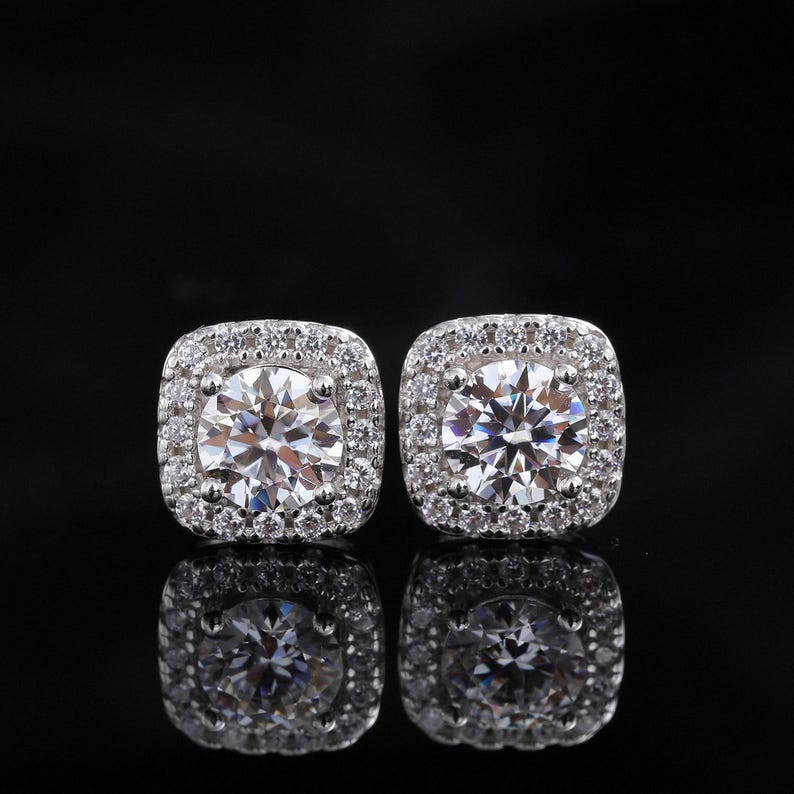 May include: A pair of square-shaped, silver-toned stud earrings. Each earring features a large, round, clear gemstone surrounded by smaller, clear gemstones. The earrings are set against a black background, with their reflections visible.
