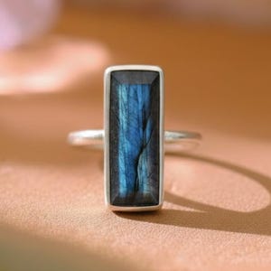 Pode incluir: Anel de prata com uma pedra labradorita azul retangular. A pedra preciosa tem listras azuis escuras e claras. O anel está em um fundo neutro, destacando a joia.