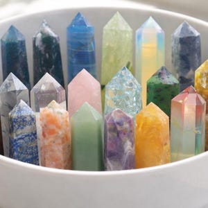 May include: A white bowl filled with a collection of colorful crystal points. The crystals vary in color, including purple, blue, green, pink, and orange. The crystals are arranged upright, showcasing their pointed tips.