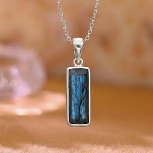 May include: A silver necklace with a rectangular pendant. The pendant features a blue gemstone with dark blue veining. The necklace is displayed on a light brown surface.