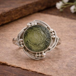 Może przedstawiać: Srebrny pierścionek z dużym, okrągłym, oliwkowozielonym kamieniem szlachetnym. Pierścionek ma dekoracyjny, wirujący wzór wokół kamienia, z małymi srebrnymi koralikami. Pierścionek leży na brązowej powierzchni.