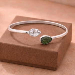 May include: A silver bangle bracelet with an open design. One end features a clear, faceted gemstone, the other a green, teardrop-shaped stone. The bracelet is displayed on a brown surface.