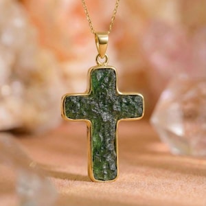May include: A gold-colored pendant necklace featuring a cross-shaped pendant. The cross has a textured, green stone center and is framed by a gold-colored border. The necklace chain is also gold-colored.