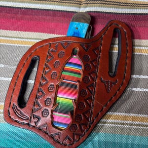 Puede incluir: Una funda de cuchillo de cuero marrón con un diseño de serape colorido. La funda tiene un recorte en forma de botella con un patrón de serape a rayas de arcoíris. El mango del cuchillo es azul y la funda tiene patrones florales y geométricos.