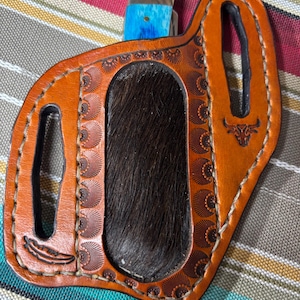 Puede incluir: Una funda de cuchillo de cuero marrón con un inserto de piel marrón oscuro. La funda tiene patrones en relieve y un borde cosido. Se ve un mango de cuchillo azul y marrón. La funda tiene un pequeño emblema de cabeza de toro.