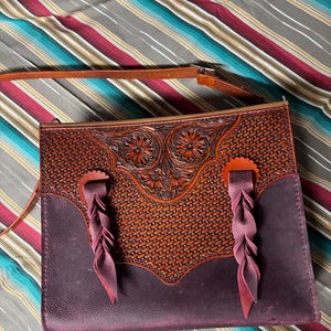 May include: A handcrafted leather purse with intricate floral and woven patterns. The purse features a rich brown and deep purple color scheme, with braided leather accents and a long strap. The bag is designed with a western aesthetic.
