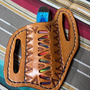 Puede incluir: Una funda de cuchillo de cuero marrón con un mango de cuchillo azul y negro. La funda tiene un patrón geométrico decorativo y costuras azules. La funda tiene ranuras con detalles coloridos.