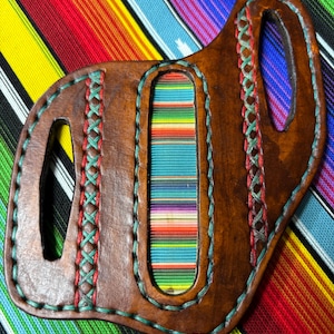 Puede incluir: Funda de cuero cosida a mano con un inserto de tela a rayas coloridas. La funda es marrón con costuras turquesa y rojas. El diseño incluye formas recortadas y un asa. El fondo presenta un textil vibrante a rayas.