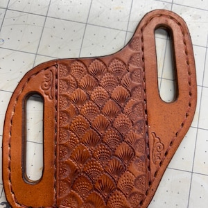 Puede incluir: Una funda de cuero marrón hecha a mano con un patrón en relieve detallado. La funda presenta un diseño decorativo en forma de abanico y dos ranuras para un cinturón. Los bordes están cosidos con un hilo en contraste.