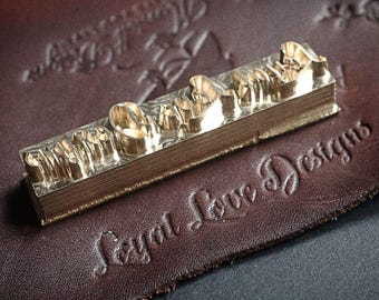 May include: A brass stamp with the text "Loyal Love Designs" resting on a piece of brown leather. The stamp is rectangular and has raised lettering. The leather has embossed text and a circular logo.