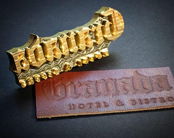 Może przedstawiać: Złoty metalowy stempel z napisem "Brampton" w gotyckiej czcionce. Brązowa skórzana etykieta z wytłoczonym tekstem "Brampton HOTEL & BISTRO" znajduje się obok stempla. Tło jest ciemne.