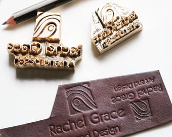 May include: A close-up shot of a branding design setup. The image features two wooden stamps, a leather tag, and a pencil. The stamps and tag display the "Rachel Grace Art and Design" logo. The leather tag is a deep burgundy color.
