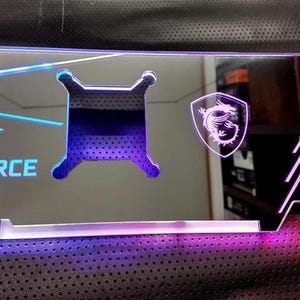 Könnte beinhalten: Eine transparente GeForce RTX-Komponente aus Acryl mit blauer und violetter LED-Beleuchtung. Das Design umfasst das MSI-Logo und ein stilisiertes Kühlelement. Der Text "GEFORCE RTX" ist in Blau sichtbar.
