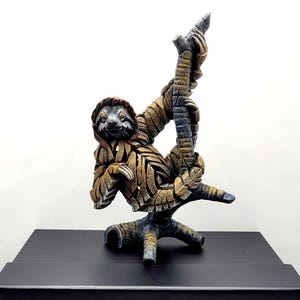 May include: A sloth sculpture with a textured, layered brown and tan finish, hanging from a dark branch. The sculpture is positioned on a black rectangular box featuring the white text EDGE SCULPTURE MINIATURE printed on the front face.