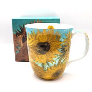 Può includere: Una tazza in ceramica con una vivace stampa di girasoli su sfondo azzurro, ispirata all'opera di Van Gogh. La tazza presenta un interno bianco ed è posizionata davanti a una scatola rettangolare coordinata con lo stesso motivo floreale.