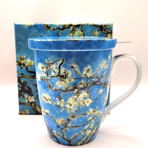 May include: A ceramic mug with a lid featuring a blue background and white almond blossom branches. The mug has a curved handle and is positioned in front of a matching square box that displays the same floral pattern in shades of blue and white.