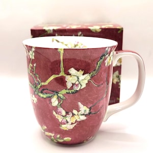 Può includere: Una tazza in ceramica con uno sfondo bordeaux intenso caratterizzato da un motivo dipinto di fiori di mandorlo chiari su rami marroni. L'interno della tazza è bianco e include una scatola bordeaux abbinata con lo stesso motivo floreale.