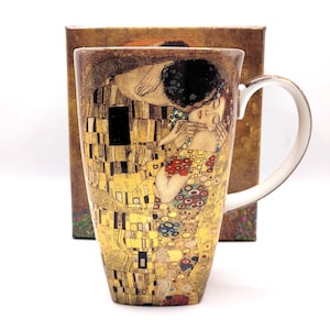 Puede incluir: Una taza alta de cerámica con una reproducción del cuadro El Beso de Gustav Klimt, que muestra a dos figuras en un abrazo con patrones dorados, negros y florales. La taza tiene un interior blanco y una caja cuadrada a juego detrás.