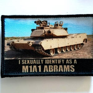 Puede incluir: Parche rectangular con un tanque M1A1 Abrams en beige y marrón, sobre un cielo azul y un paisaje desértico. El texto "I SEXUALLY IDENTIFY AS A M1A1 ABRAMS" se muestra en letras beige sobre una pancarta negra.