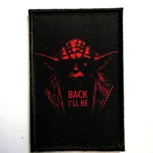 Puede incluir: Parche rectangular negro con un gráfico rojo de la cara de un personaje y el texto "BACK I'LL BE". El parche tiene un borde cosido.