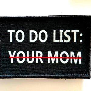 Puede incluir: Parche rectangular negro con texto blanco que dice "TO DO LIST: YOUR MOM" en una fuente en negrita, sans-serif. El texto "YOUR" está tachado con una línea roja. El parche tiene un borde negro.